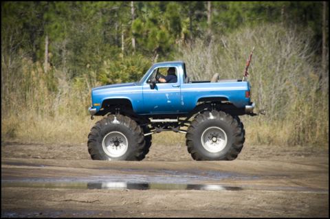 1996 Ford F-150 - A lifted blue pickup truck with massive tires navigates a muddy track, kicking up dirt.