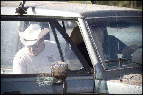 1996 Ford F-150 - Two men inside a mud-splattered vehicle, one wearing a cowboy hat and the other at the wheel.