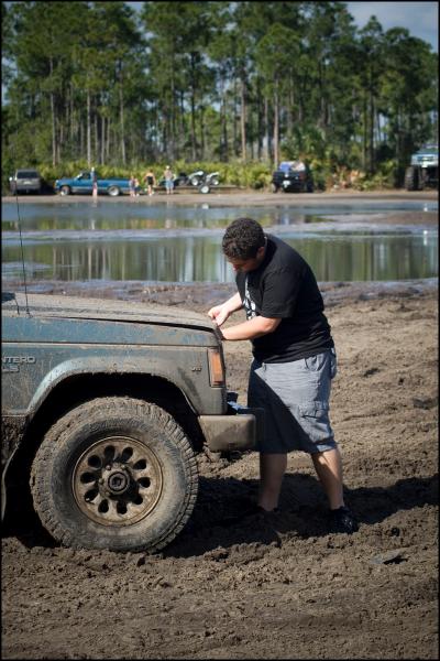 1996 Ford F-150 - A person inspects the engine of a muddy blue SUV stuck in a mire, with other vehicles and trees in the background.
