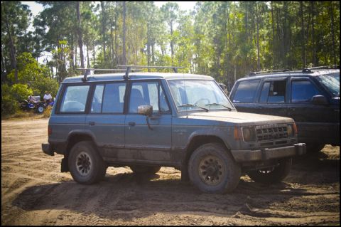 1996 Ford F-150 - A blue Mitsubishi Montero is parked on a dirt road, with a dark SUV behind it and trees in the background.