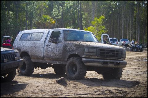 1996 Ford F-150 - A muddy 1996 Ford F-150 pickup truck with a camper shell is seen off-road, with other vehicles in the background.