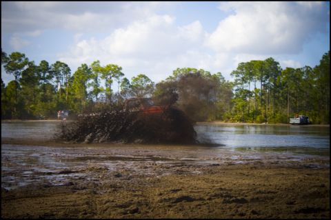 1996 Ford F-150 - An off-road vehicle navigates a muddy terrain, creating a large splash. Another vehicle is seen in the distance.