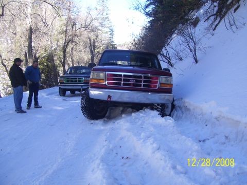 1993 Ford Bronco - Two trucks are on a snowy road with trees on either side. The front truck is partially stuck in a snowbank.
