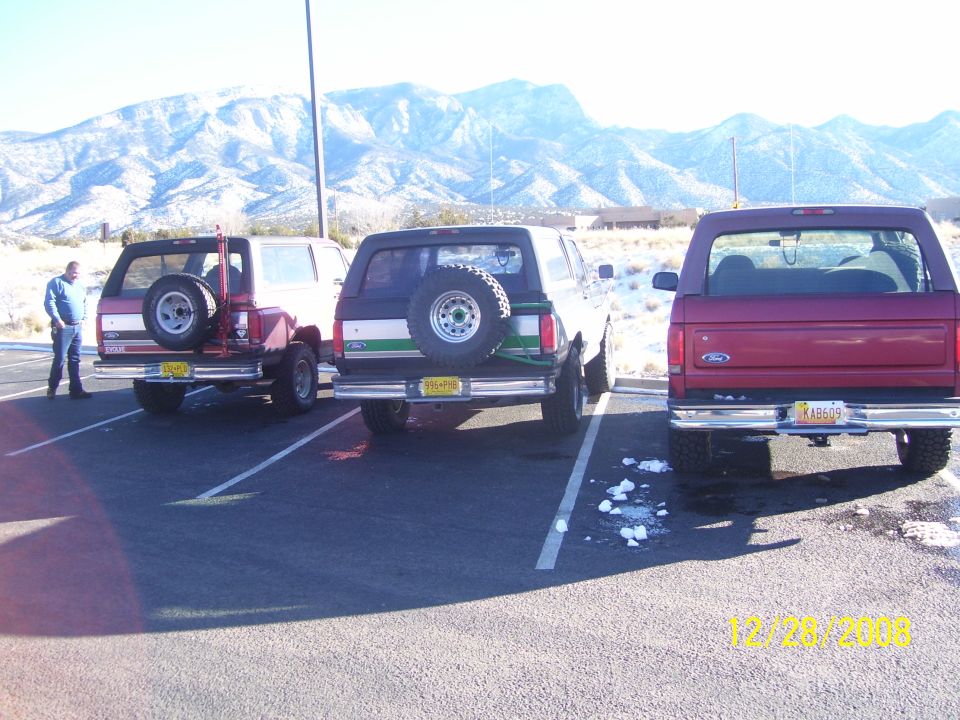 Three SUVs parked in a lot with mountains in the background. One is red, one is white with green stripes, and one is red with a spare tire on the back