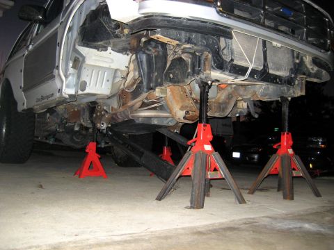 1996 Ford Bronco - The front suspension of a silver truck is elevated on red jack stands, revealing a heavily rusted frame and grille.