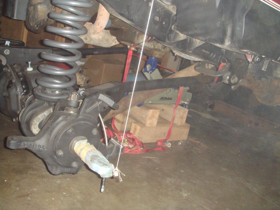 Close-up of a vehicle's front axle assembly with coil spring and brake components. Wood blocks and straps are visible
