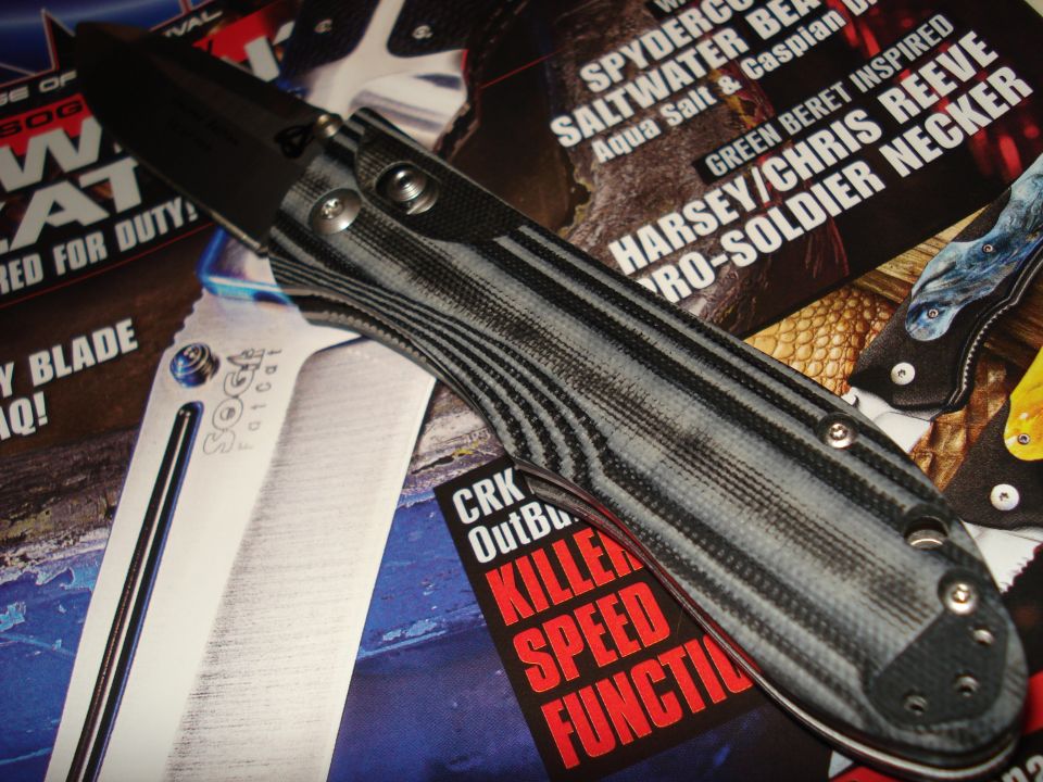 Close-up of a folding knife with a gray and black striped handle, resting on magazine pages with knife advertisements