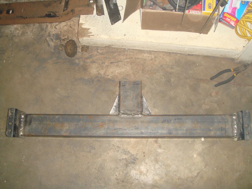 Fabricated steel crossmember with central support and end brackets on concrete floor.