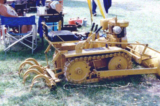 Yellow tracked tiller with curved tines on front. Engine visible on top