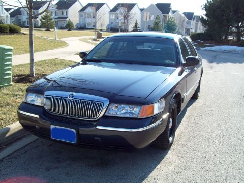 1999 Mercury Grand Marquis - A dark-colored Mercury Grand Marquis is parked in front of a row of suburban houses. The front of the car, including the grille and headlig…