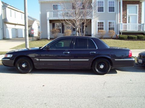 1999 Mercury Grand Marquis - Full driver-side profile of a dark blue Mercury Grand Marquis parked at the curb of a suburban townhome street. The sedan sits on black ste…