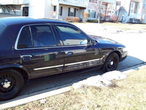 1999 Mercury Grand Marquis - The side profile of a dark blue 1999 Mercury Grand Marquis is shown parked next to a curb with patchy snow on the grass. The car features c…