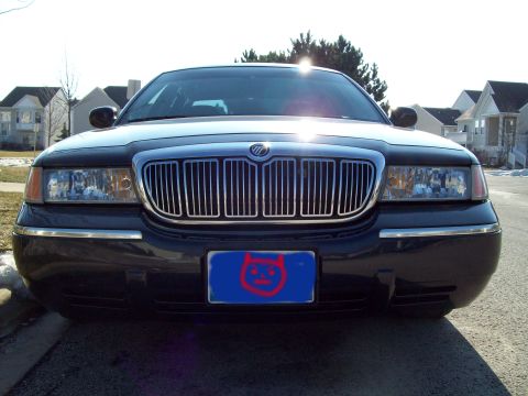 1999 Mercury Grand Marquis - Front view of a dark colored sedan with a chrome grille and headlights. A blue license plate with a red cartoon devil face is mounted on th…