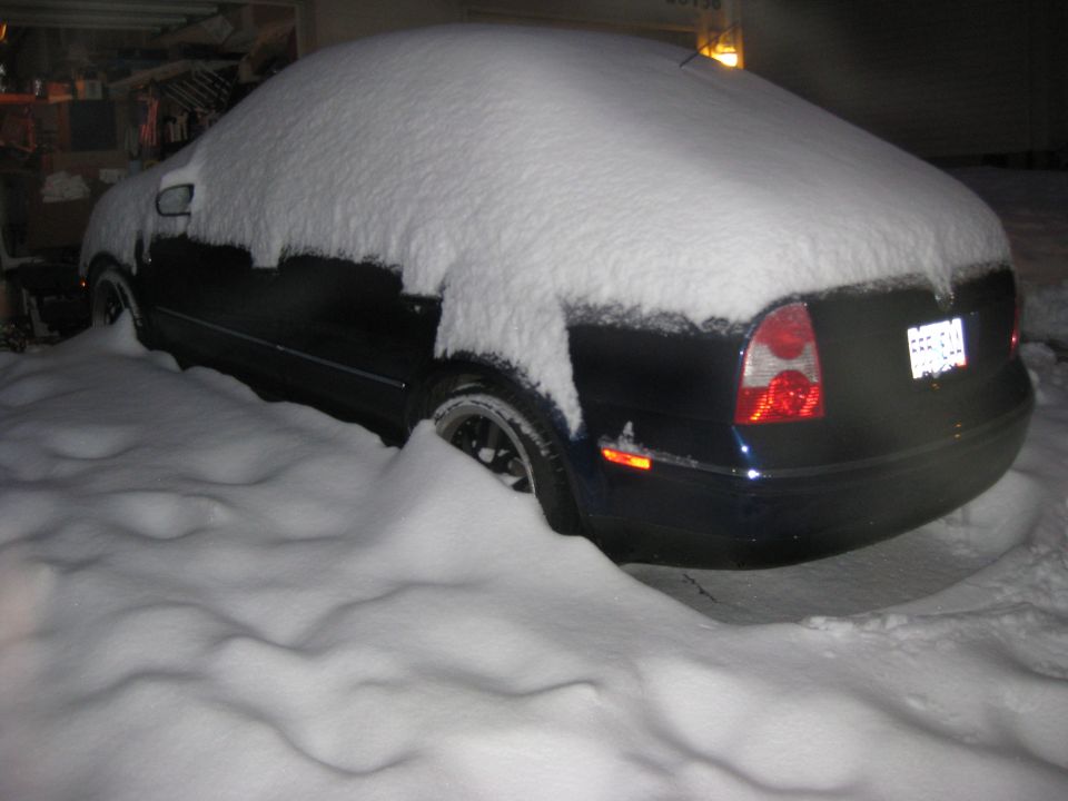 Dark blue VW Passat sedan covered in deep snow at night, rear three-quarter view