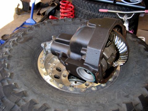1996 Ford Bronco - A differential housing with visible gears rests on a large off-road tire. In the background, a red coil spring and a blue jack are visible.