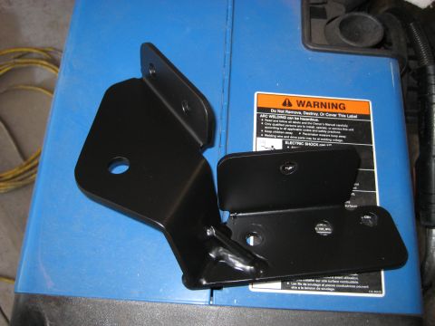 1996 Ford Bronco - Two black metal brackets are placed on top of a blue surface, which appears to be part of a welding machine. A warning label with orange an…