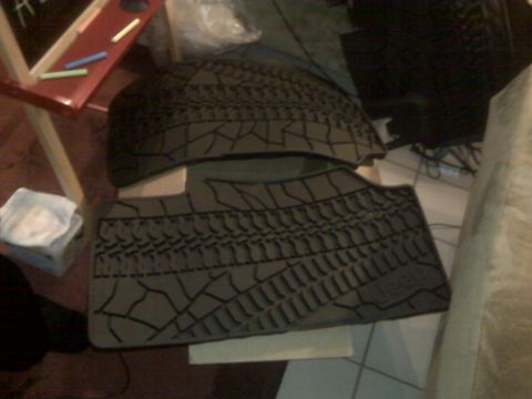 2008 Jeep Wrangler - Two dark grey Jeep floor mats with a tire tread pattern are laid out on a tiled floor. The mats have a textured surface with a cracked eart…