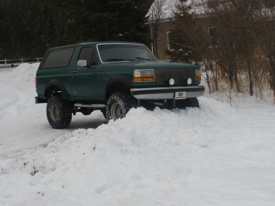 Green lifted two-door Ford Bronco on large mud tires parked in snow, front three-quarter view.