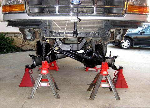 1996 Ford Bronco - The underside of a Ford Bronco is supported by multiple red jack stands. The front grille and headlights are visible above the suspension c…