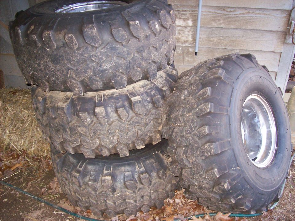 Stack of four used mud-terrain tires on chrome wheels leaning against a wood shed