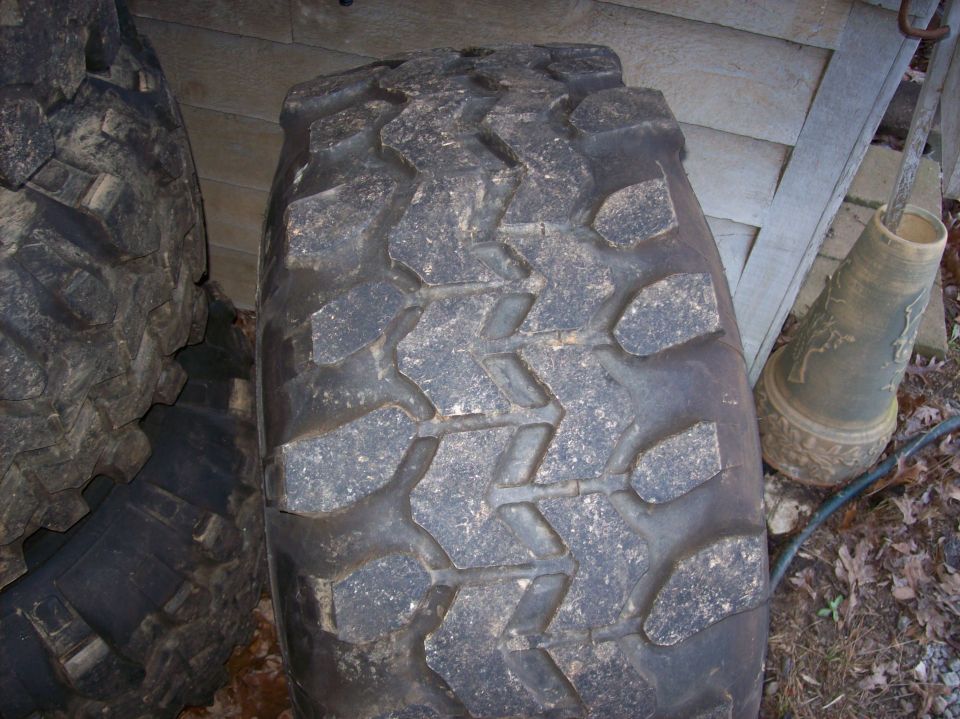 Upright wide mud-terrain tire showing chunky tread face against a wooden shed
