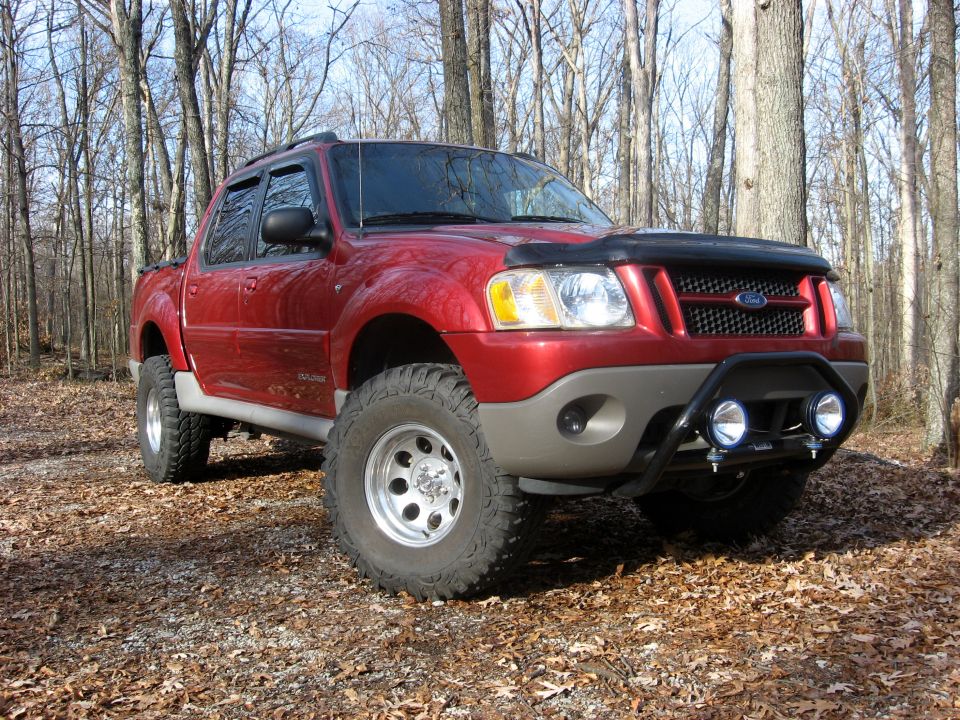 Red Ford Sport Trac pickup truck with off-road tires and brush guard in a forest.