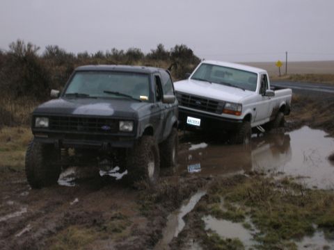 1984 Ford Bronco II - Two off-road vehicles are stuck in a muddy field under an overcast sky.