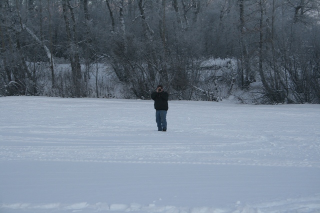 Person stands in a vast snowy field, trees in the background