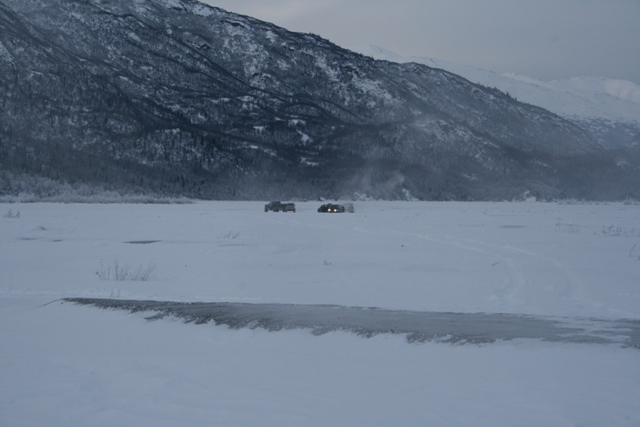 Two boxy vehicles with headlights on, parked in a vast, snow-covered plain with a forested, snow-capped mountain range in the background