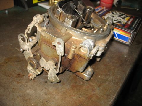 1978 Ford Bronco - A carburetor sits on a workbench, showing signs of wear and rust, with a box of automotive parts nearby.