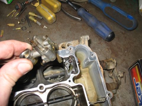 1978 Ford Bronco - A hand holds a carburetor part over a disassembled carburetor, with various tools and small parts scattered on the surface.