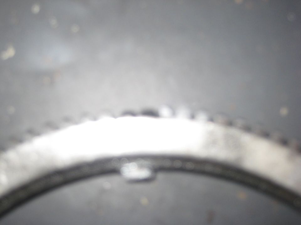Blurry close-up of a toothed metal ring gear, possibly a flywheel/flexplate ring gear, on a dark surface