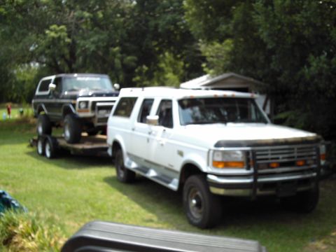 1978 Ford Bronco - White Ford F-Series truck tows a dark Ford Bronco on a trailer in a grassy area.