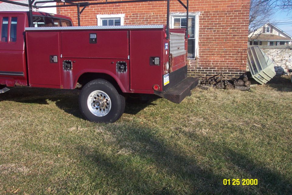 Maroon Ford F-250 pickup truck with utility body, parked on grass next to a brick building.