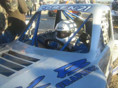 1990 Ford Bronco - Photograph of a helmeted driver in the roll-caged cockpit of a blue off-road race truck, not a technical document.