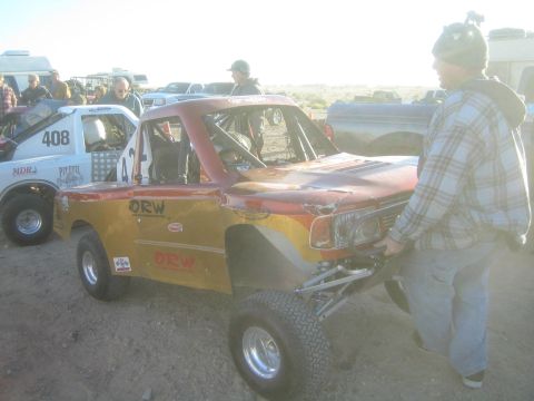 1990 Ford Bronco - Photograph of an orange off-road race truck being maneuvered by hand in a desert pit area, not a technical document.