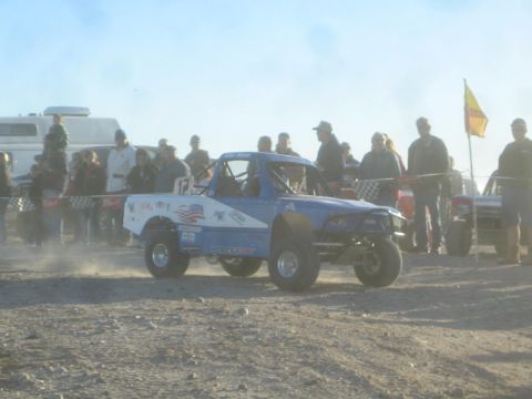 1990 Ford Bronco - Photograph of a blue and white off-road race truck on a dusty course with spectators, not a technical document.