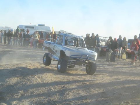 1990 Ford Bronco - Photograph of a blue off-road race truck catching air on a dirt course near a crowd, not a technical document.