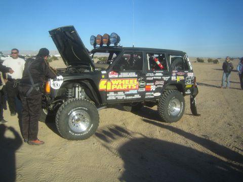 1990 Ford Bronco - Photograph of a black sponsor-decaled Jeep Cherokee race rig with hood raised in the desert pits, not a technical document.
