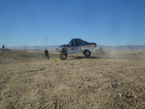 1990 Ford Bronco - Photograph of a blue off-road race truck catching air on a desert course near power lines, not a technical document.
