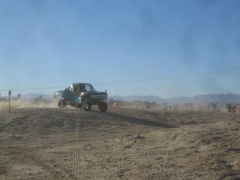 1990 Ford Bronco - Photograph of a teal off-road race truck catching air over a desert berm, not a technical document.