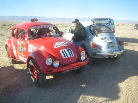 1990 Ford Bronco - red number 1117 race-prepped VW Beetle and a silver Beetle parked nose-to-tail on a dirt road as a man stands between them.