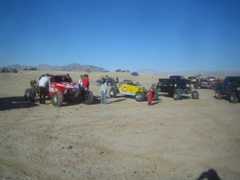 1990 Ford Bronco - lineup of off-road race trucks and buggies in red, yellow and black staged across a flat desert lot with people gathered around.