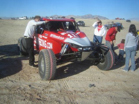 1990 Ford Bronco - red and silver tube-chassis off-road race buggy sits on dirt as spectators and a man leaning on it stand nearby under desert hills.