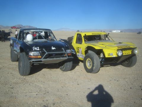 1990 Ford Bronco - black number 742 off-road prerunner and a yellow number 15 race truck parked side by side on a flat desert lot.
