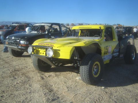 1990 Ford Bronco - yellow number 15 off-road race truck with tubular fenders and round lights staged on dirt among other race vehicles.