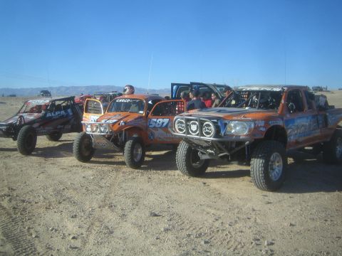 1990 Ford Bronco - Several orange and red off-road prerunner trucks with roof light bars and round driving lights lined up on dirt under a blue sky.