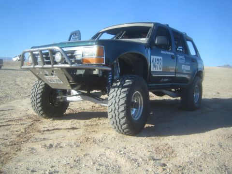1990 Ford Bronco - lifted dark green SUV marked 1478 with a tubular front bumper and large mud tires sits on a dirt lot under a clear sky.