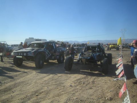 1990 Ford Bronco - Off-road race trucks and a buggy staged on a dusty desert lot among a crowd, with checkered course flags strung in the foreground.