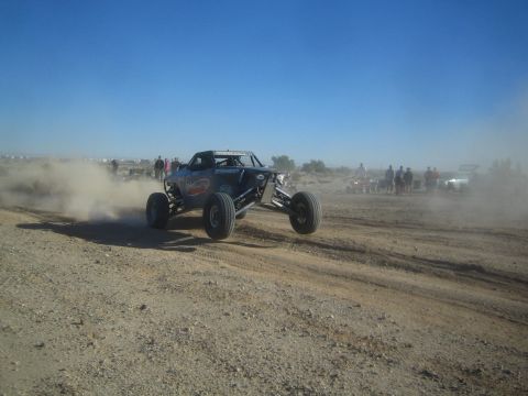 1990 Ford Bronco - blue tube-chassis off-road buggy corners hard and throws a cloud of dust on a desert track lined with spectators.